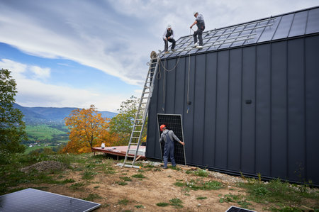 Men mounters installing solar panel system on roof of house. Workers in helmets lifting up photovoltaic solar module with help of ropes outdoors. Concept of alternative and renewable energy.の写真素材