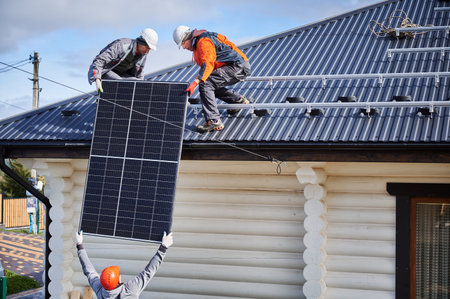 Men builders installing solar panel system on roof of house. Electricians in helmets lifting up photovoltaic solar module outdoors. Concept of alternative and renewable energy.の写真素材