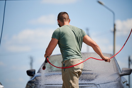 Confident man washing his car on self car-washing station. Cleaning car using active foam. Back view of young man cleaning his auto by water gun.の写真素材