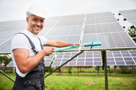 Professional worker cleaning solar panel from dust. Smiling man wiping PV battery using mop. Man holding mop and smiling straight to camera.の写真素材