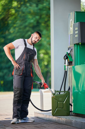 Professional worker refueling canister with fuel. Portrait of skilled man filling cistern with gasoline for customer. Man in ovealls with pump nozzle refueling canister next to gasoline column.の写真素材