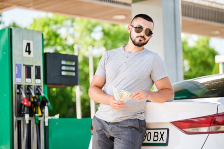 Bearded man waiting for petrol station operator to pay for fuel. Man leaning on his car and counting how much gasoline costs. Male in casual clothes holding money and looking straight into camera.の写真素材