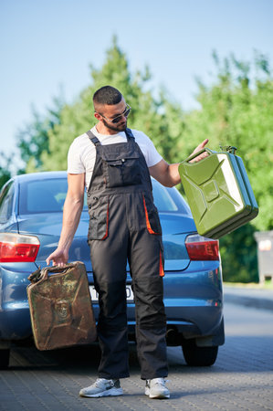 Gas station worker refueled two canisters and waiting for customer. Young operator holding cisterns full of gasoline. Male in overalls with canisters in both hands on background of blue car.の写真素材