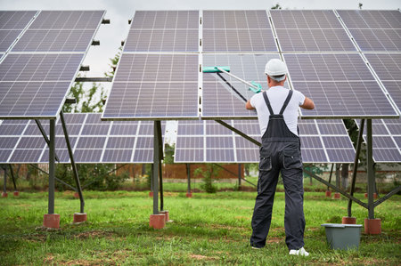 Technician cleaning solar photovoltaic panels by mop. Back view of man in overalls and helmet taking care about PV battery. Alternative energy innovations concept.の写真素材