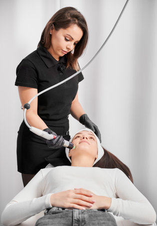 Medical esthetician cosmetologist using radiofrequency device while performing rejuvenation facial treatment in cosmetology clinic. Woman having skincare face lifting procedure in beauty salon.の写真素材