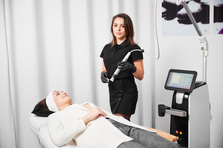 Female cosmetologist holding radiofrequency device and looking at camera while standing by client in cosmetology clinic. Woman receiving rejuvenation skincare treatment in beauty salon.の写真素材