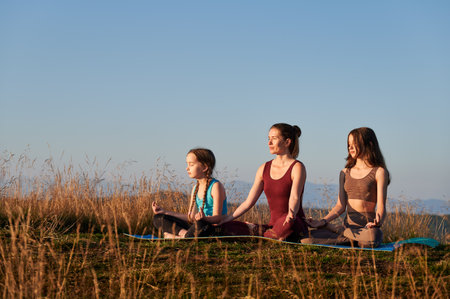 Young mother meditating with her daughters. Meditation at dawn in mountains. Three female sitting in lotus position at early morning.の写真素材