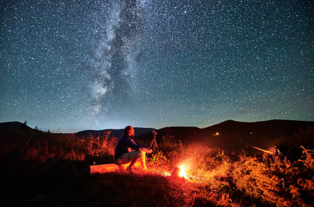 Man taking photos of starry sky with Milky way. Male photographer sitting on log and focusing camera. Man with camera sitting next to camp fire in mountains.の写真素材