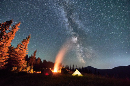 Night camping under sky full of stars and Milky way in the mountains. Starry sky over illuminated tourist tents on hills near forest. Warm light from campfire at dark night.の写真素材
