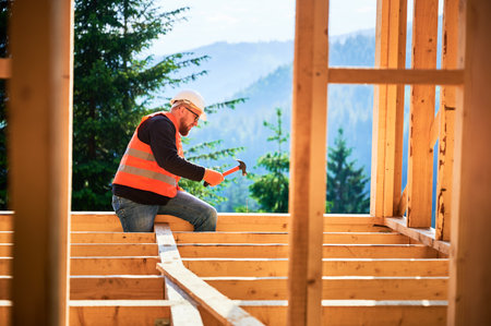 Carpenter constructing two-story wooden frame house near forest. Man hammering nails into the structure, wearing protective helmet and construction vest. Concept of modern ecological construction.の写真素材