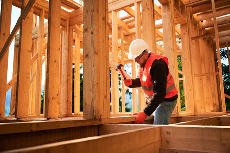Carpenter constructing wooden frame house. Bearded man with glasses is hammering nails into the structure, wearing protective helmet and construction vest. Concept of modern ecological construction.の写真素材