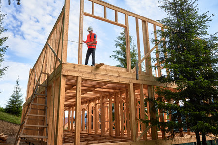 Carpenter constructing timber-framed two-story building. Man measures distances with measuring tape while dressed in workwear and hard hat. Ideology of contemporary, eco-friendly building practices.の写真素材