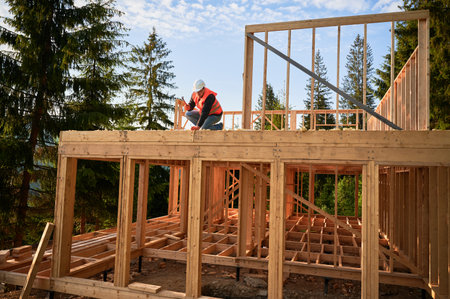 Carpenter constructing two-story wooden frame house near forest. Bearded man hammering nails into the structure, wearing protective helmet, construction vest. Concept of modern ecological constructionの写真素材