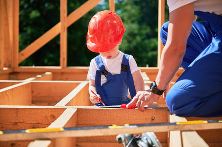 Father with toddler son constructing wooden frame house. Male builder playing with kid on construction site, wearing helmets and blue overalls on sunny day. Carpentry and family concept.の写真素材