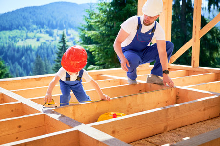 Father with toddler son building wooden frame house. Man worker instructing his son on measurement of distance using tape measure on construction site. Carpentry and family concept.の写真素材