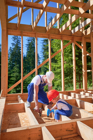 Father with toddler son building wooden frame house. Male builder educating his son on the utilization of power screwdriver on construction site on sunny day. Carpentry and family concept.の写真素材
