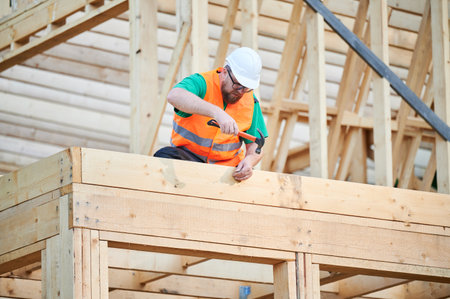 Carpenter constructing two-story wooden frame house. Man wearing glasses hammering nails into structure while wearing protective helmet and construction vest. Concept of modern ecological constructionの写真素材