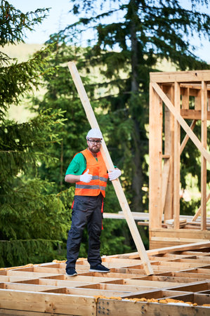 Carpenter builds wooden frame house near the forest. Bearded man with spectacles holds large plank, showing thumbs up, wearing work clothes and helmet. Concept of modern ecological construction.の写真素材