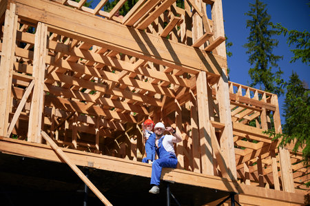 Father with toddler son building wooden frame house. Man showing his son the view from future window at construction site, wearing helmets and blue overalls on sunny day. Carpentry and family concept.の写真素材