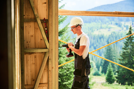 Carpenter constructing wooden framed house. Man worker in glasses working with screwdriver, wearing work overalls and helmet. Concept of modern eco-friendly construction.の写真素材