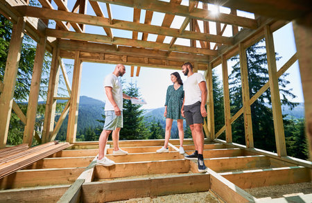 Man with plan showcases to couple the process of constructing wooden framed house. Investors inspecting their future dwelling in mountains near forest. Concept of contemporary ecological construction.の写真素材
