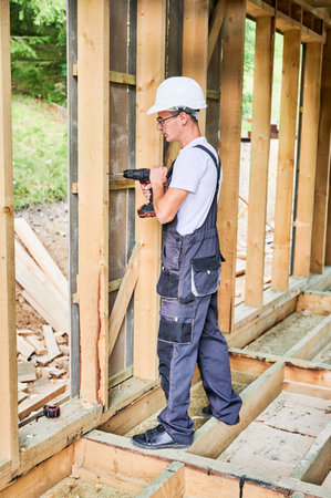 Carpenter constructing wooden framed house. Man worker in glasses working with screwdriver, wearing work overalls and helmet. Concept of modern eco-friendly construction.の写真素材