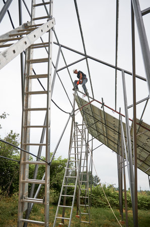 Solar installer standing on metal rail and building photovoltaic solar panel system under white sky. Male worker assembling supporting structure for solar modules.の写真素材