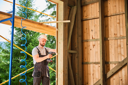 Carpenter constructing wooden framed house. Bearded man worker cladding facade of house, fastening with screwdriver, wearing work overalls and helmet. Concept of modern eco-friendly construction.の写真素材