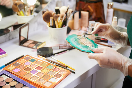 Close up of female hands in sterile gloves blending skin foundation. Woman makeup artist standing by dressing table with eyeshadow palette and preparing foundation for professional makeup.の写真素材