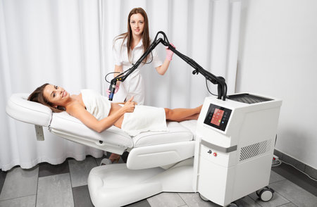 Smiling client lying on white couch and showing class during procedure of abdomen skin laser resurfacing in beauty salon. Beautiful young woman satisfied with skin rejuvenation procedure.の写真素材