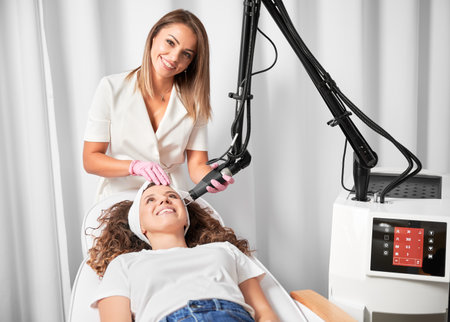 Female doctor cosmetologist using erbium ablative laser machine while performing resurfacing procedure. Smiling woman lying on daybed and receiving facial skincare treatment in beauty salon.の写真素材