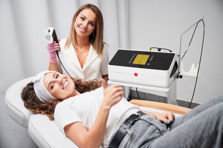 Portrait of happy cosmetologist and young woman patient showing approval gesture after laser skincare procedure. Beautician holding laser scanner device for skin resurfacing treatment.の写真素材