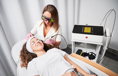 Woman cosmetologist painlessly and quickly removing telangiectasias from face. Happy patient with protection for eyes who undergoing laser treatment of facial vessels in beauty salon.の写真素材