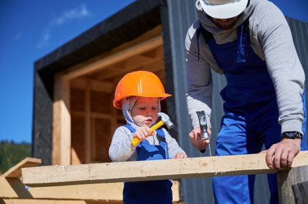 Father with toddler son building wooden frame house. Boy helping his daddy, hammering nail into plank on construction site, wearing helmet and blue overalls on sunny day. Carpentry and family concept.の写真素材