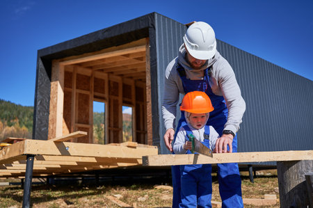 Father with toddler son building wooden frame house on pile foundation. Male builders using hand saw to cut boards on construction site on sunny day. Carpentry and family concept.の写真素材