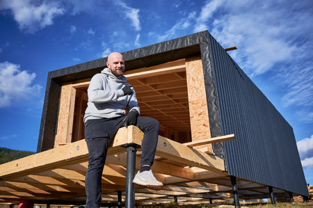 Male inspector building wooden frame house on pile foundation. Portrait of bald smiling man sitting on terrace on construction site, inspecting quality of work.の写真素材