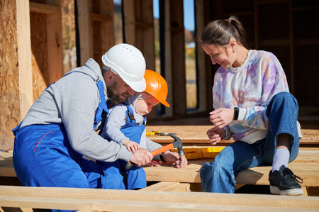 Father, mother and son building wooden frame house. Toddler boy helping his daddy, while woman looking for them on construction site. Guys wearing helmet and blue overalls. Carpentry, family concept.の写真素材