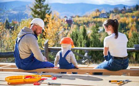 Back view of father, mother and son building wooden frame house. Toddler boy, daddy and woman having a rest on construction site. Guys wearing helmet and blue overalls. Carpentry, family concept.の写真素材