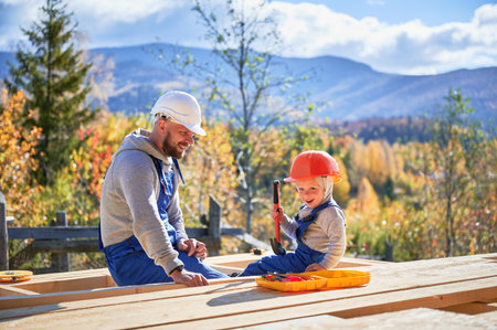 Father with toddler son building wooden frame house. Male builders hammering nail into plank on construction site, wearing helmet and blue overalls on sunny day. Carpentry and family concept.の写真素材
