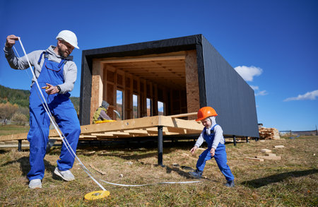 Father with toddler son building wooden frame house in the Scandinavian style barnhouse. Male builder and kid playing with tape measure on construction site on sunny day. Carpentry and family concept.の写真素材