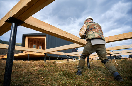 Man building wooden house on pile foundation. Back view of male worker drilling hole by electric...