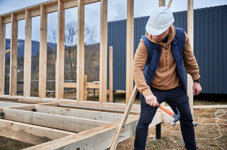 Man worker building wooden frame house on pile foundation. Carpenter hammering nail into wooden plank, using hammer. Carpentry concept.の写真素材