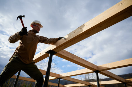 Man worker building wooden frame house on pile foundation. Carpenter hammering bolt into wooden joist, using hammer. Carpentry concept.の写真素材