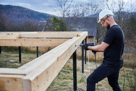 Man building wooden frame house on pile foundation. Male worker drilling hole by drill in timber framing of future house. Builder wearing white helmet and protective goggles. Carpentry concept.の写真素材
