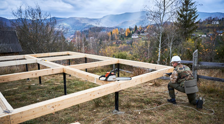 Man building frame house on pile foundation. Male worker drilling hole by electric drill in wooden frame of future house on construction site. Carpentry concept.の写真素材