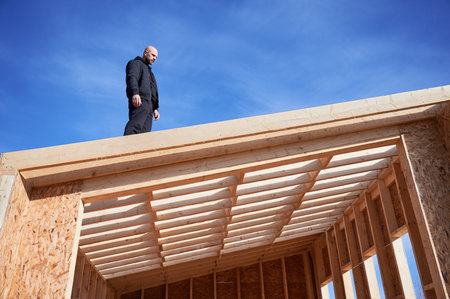 Male designer building wooden frame house. Bald man standing on construction site, inspecting quality of work on sunny day with blue sky on background.の写真素材