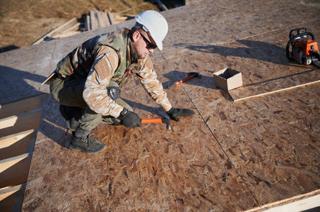 Carpenter hammering nail into OSB panel on the roof top of future cottage. Man worker building wooden frame house. Carpentry and construction concept.の写真素材