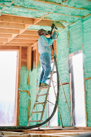 Male builder insulating wooden frame house. Man worker spraying polyurethane foam inside of future cottage, standing on ladder, using plural component gun. Construction and insulation concept.の写真素材