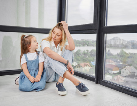 Woman and little girl sitting on the floor and looking to each other in family new home. Mother and child enjoying view from panoramic window in newly built apartment.の写真素材
