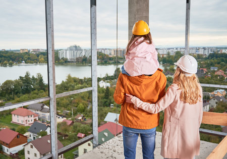 Back view of parents with daughter standing inside apartment building under construction. Man and woman with child wearing building helmets while enjoying city view from future home.の写真素材